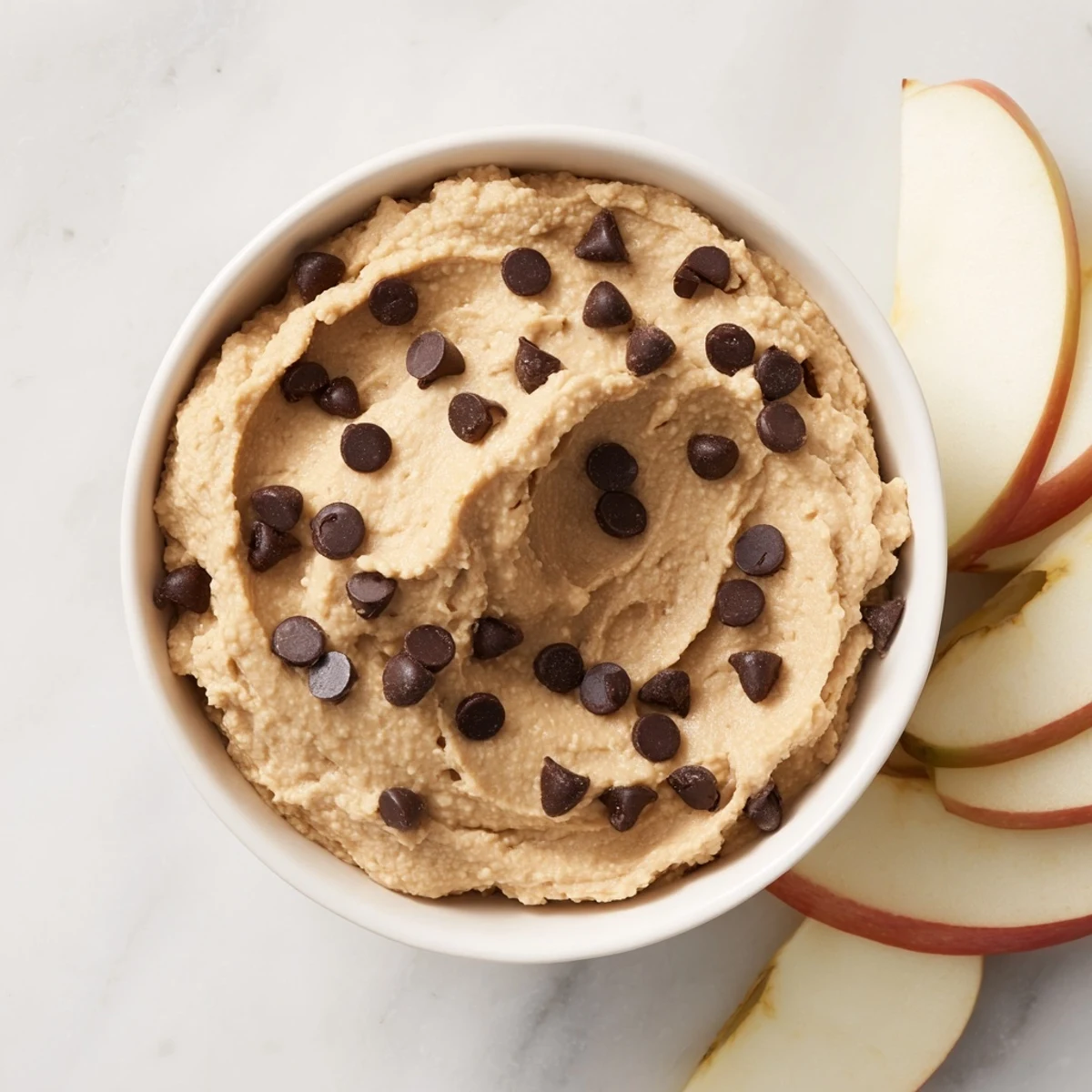 Creamy chocolate chip cookie dough hummus scooped into a bowl next to crisp apple slices on a platter for dipping.