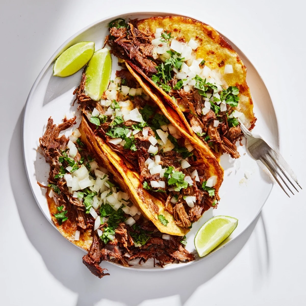 Slow cooker birria tacos dipped in rich red consommé beside lime wedges.