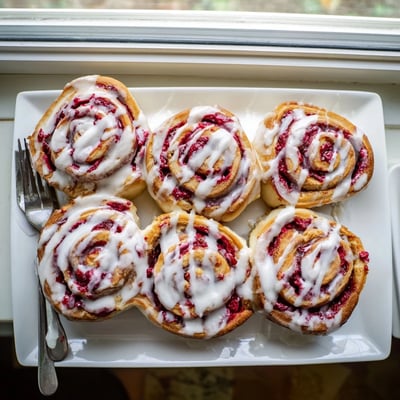 A warm stack of Raspberry Cinnamon Rolls With Lemon Glaze is drizzled with creamy frosting, perfect for a cozy brunch with a cup of coffee.